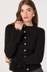 A young woman is wearing a black textured cardigan with a fuzzy trim on the cuffs and around the button placket. The cardigan has a row of silver buttons down the front. She has long brown hair, light blue eyes, and is accessorized with silver rings on her fingers and a white rectangular earring.